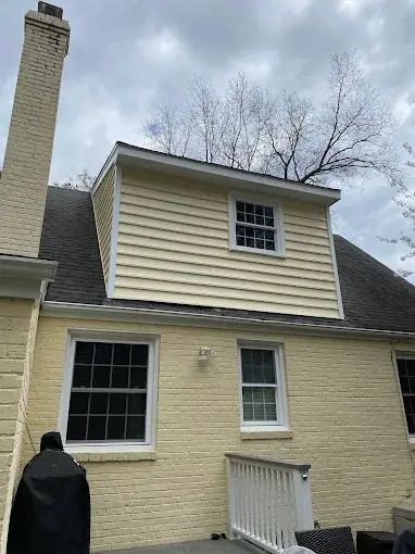 The completed exterior of a second-story home addition featuring pale yellow horizontal siding and a new white-framed window, built above a painted brick ground floor.