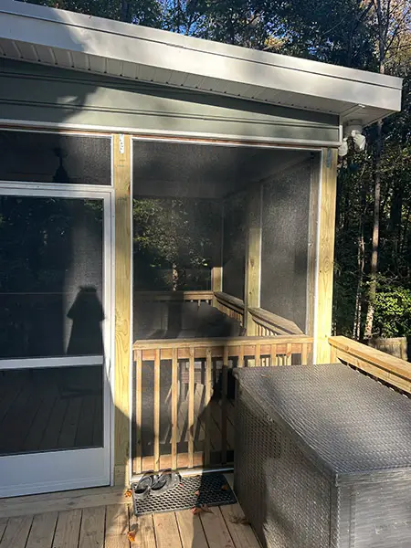 An exterior view of a screened-in porch addition from the deck level, featuring a wooden frame, black mesh screens, a white sliding screen door, and a large grey wicker storage box on the wooden floor.