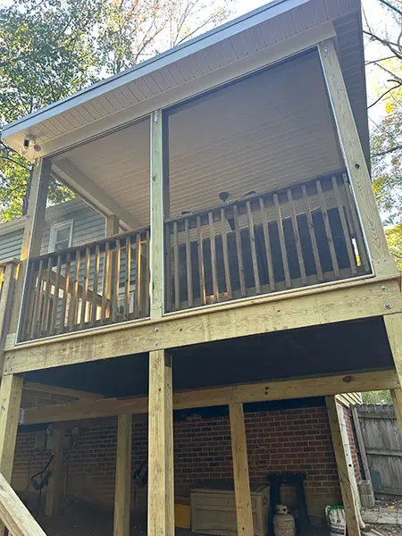 A ground-level perspective looking up at an elevated screened-in porch supported by thick wooden posts and beams, showing the open space beneath the structure against a brick house wall.