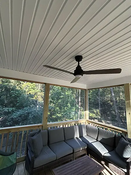 An interior view of a screened-in porch featuring a large grey L-shaped sectional sofa, a white paneled ceiling with a dark ceiling fan, and wooden railings overlooking a lush green wooded area.