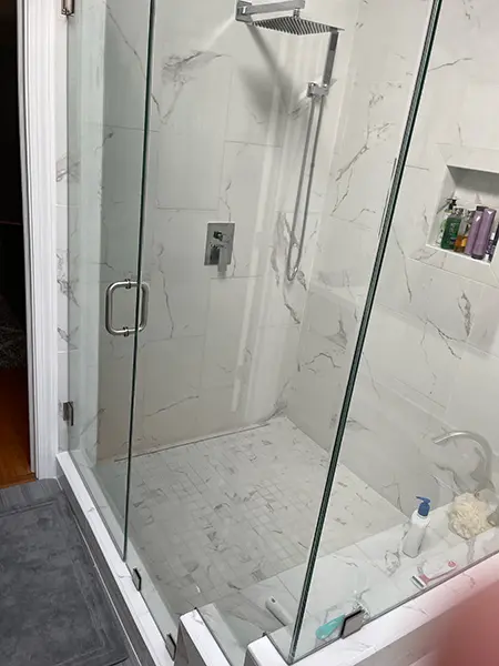 A modern walk-in shower with glass doors, featuring large white marble-patterned tiles on the walls and small white mosaic floor tiles. It is equipped with a square rain shower head, a handheld sprayer, and a built-in wall niche for toiletries.