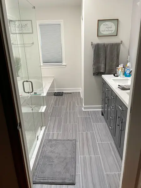 A view of a bathroom featuring grey wood-look floor tiles and a long grey double vanity with a white countertop. To the left is a glass-enclosed shower, and the back wall has a window with white blinds and light grey walls.