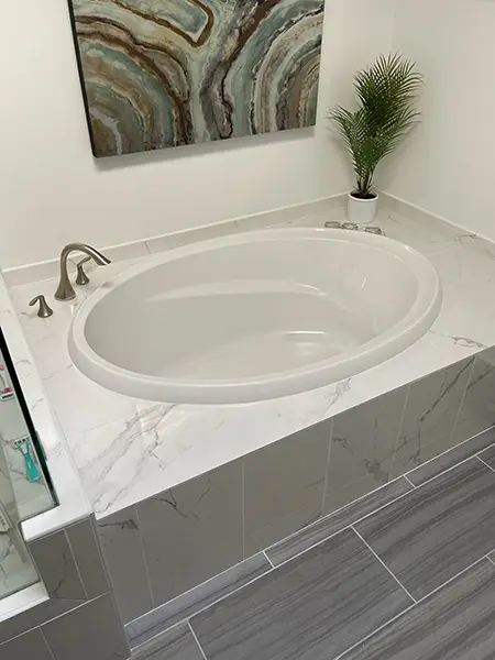 An oval white soaking tub surrounded by white marble-style tiles. The area includes brushed nickel fixtures, an abstract green and gold painting on the wall, and a small green plant in a white pot.