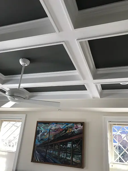 A low-angle view of a white coffered ceiling with dark gray recessed panels and a white ceiling fan. On the beige wall below, a vibrant painting of a train on a bridge hangs between two windows with decorative diamond-patterned panes.