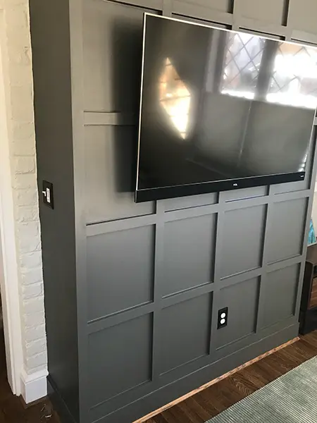 A large flat-screen TV mounted on a dark gray accent wall featuring custom rectangular grid molding. The wall is bordered by a white-painted brick column on the left and sits above dark hardwood floors.