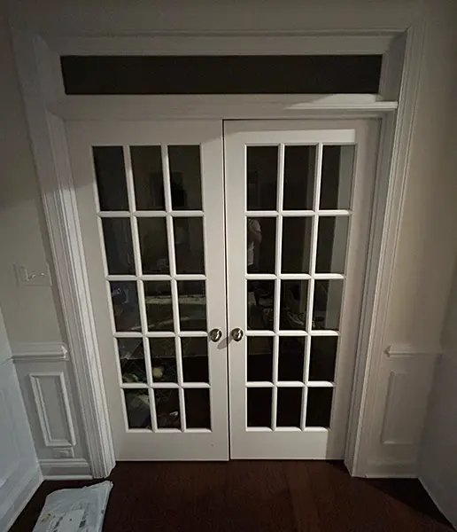 White interior French doors with a multi-pane glass grid and silver knobs, set within a decorative white frame and flanked by white wainscoted walls on dark hardwood floors.