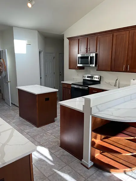 A wide view of a kitchen featuring dark wood cabinetry, white quartz countertops with grey veining, and stainless steel appliances. The space includes a small matching center island and light-colored tile flooring under a vaulted ceiling.