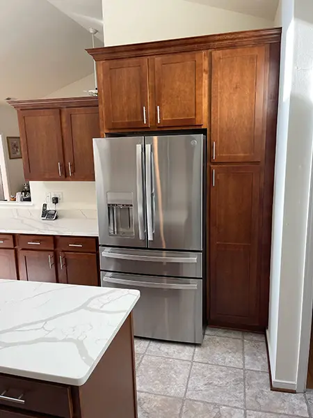 A stainless steel French door refrigerator with bottom freezer drawers, integrated into custom dark wood cabinets. The scene includes a white quartz countertop with grey veining and light-colored square floor tiles.