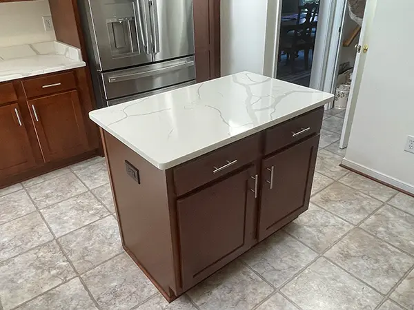 A close-up of a small kitchen island with dark wood base cabinets, silver bar handles, and a white quartz countertop with prominent grey veining, positioned on a light-colored tile floor.