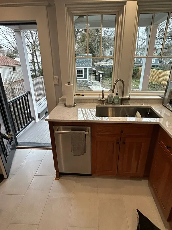 A modern kitchen interior featuring a large central marble island with grey modern stools, surrounded by medium-wood cabinets and large windows.