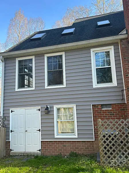 An exterior shot of a house with light grey siding and a brick base, showing multiple windows, two white utility doors, and several skylights on the black shingle roof.