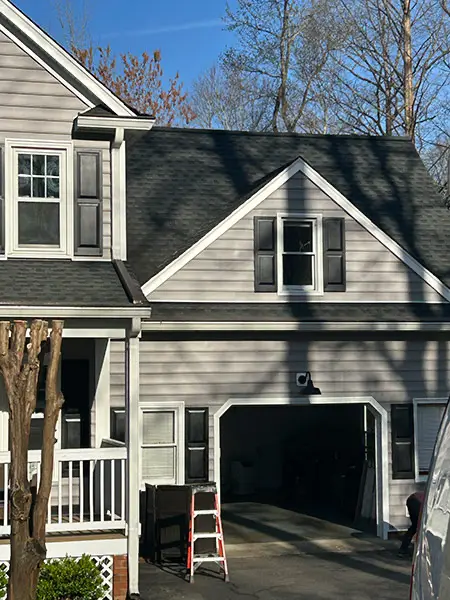 An exterior view of a home's attached garage and dormer, showing light gray siding, black shutters, and an open garage bay with a step ladder standing in the driveway.