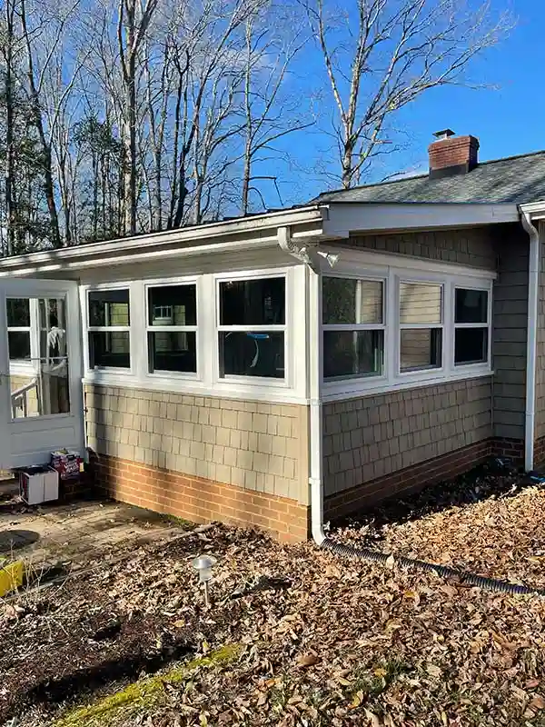 An exterior view of a home's sunroom featuring multiple large, energy-efficient white-framed windows, light beige shingle siding, and a brick patio.